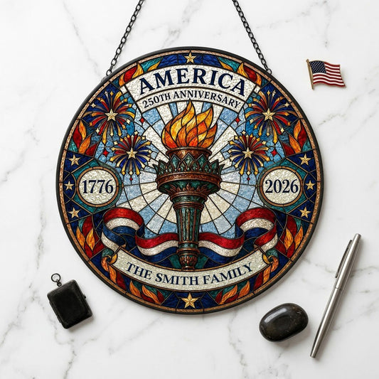 Round stained glass suncatcher with a central torch, fireworks, stars, and a red, white and blue ribbon.