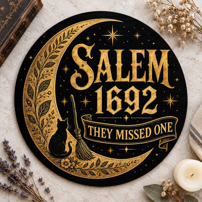 Round wooden sign with gold crescent moon, black cat, broomstick, stars and 'SALEM 1692 THEY MISSED ONE' on black.