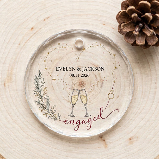 Round faceted glass ornament with gold constellation heart, ring, and clinking champagne flutes reading engaged.