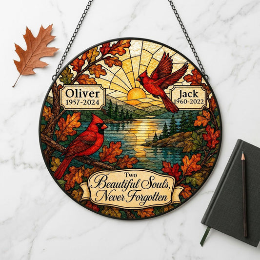 Two red cardinals with a glowing sun over a mountain lake and pine trees, framed by autumn oak leaves on a round suncatc