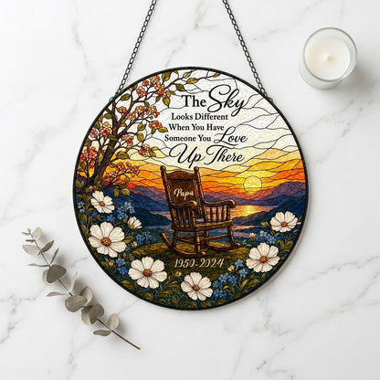 Round stained glass–style suncatcher with an empty rocking chair on a hill at sunset with flowers and a lake.