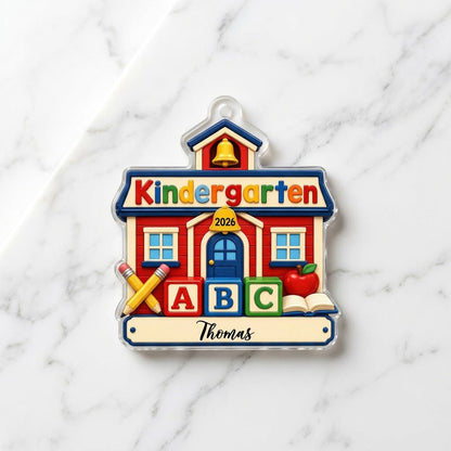 Red schoolhouse acrylic ornament with Kindergarten 2026, ABC blocks, pencils, apple, and a name on a white banner.