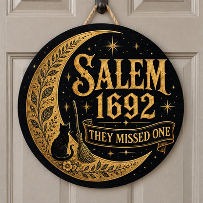 Round wooden sign with gold crescent moon, black cat, broomstick, stars and 'SALEM 1692 THEY MISSED ONE' on black.