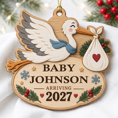 Custom-cut wood ornament of a flying stork with baby bundle above oval plaque reading BABY JOHNSON ARRIVING 2027.