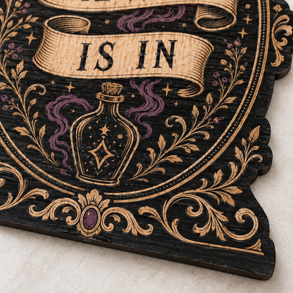 Gothic plaque sign reading "THE WITCH IS IN" with crescent moon, stars, purple smoke, potion bottle and ornate filigree 
