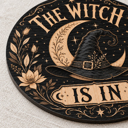 Round wooden sign with 'THE WITCH IS IN' slogan, crescent moon, witch hat, floral borders, stars and scrollwork.