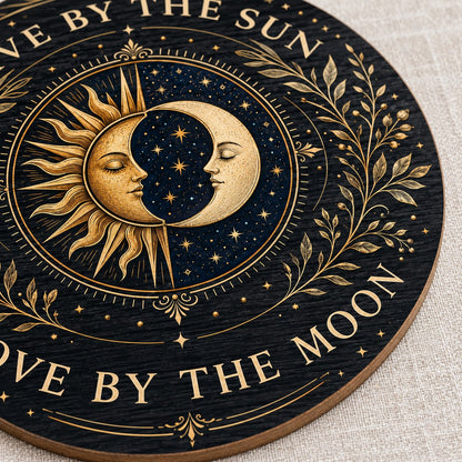 Round wooden sign with gold sun and crescent moon faces and the words “Live By The Sun, Love By The Moon”.