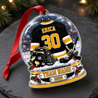 Snow globe acrylic ornament with black and gold hockey jersey, crossed sticks, puck, holly and a year plaque.