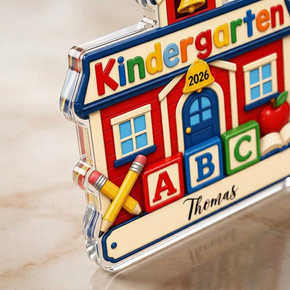 Red schoolhouse acrylic ornament with Kindergarten 2026, ABC blocks, pencils, apple, and a name on a white banner.