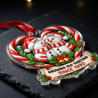 Two snowmen touching noses inside a candy-cane heart with holly, gold bells, and a scroll banner with names and year.