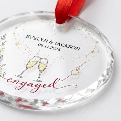 Round faceted glass ornament with gold constellation heart, ring, and clinking champagne flutes reading engaged.