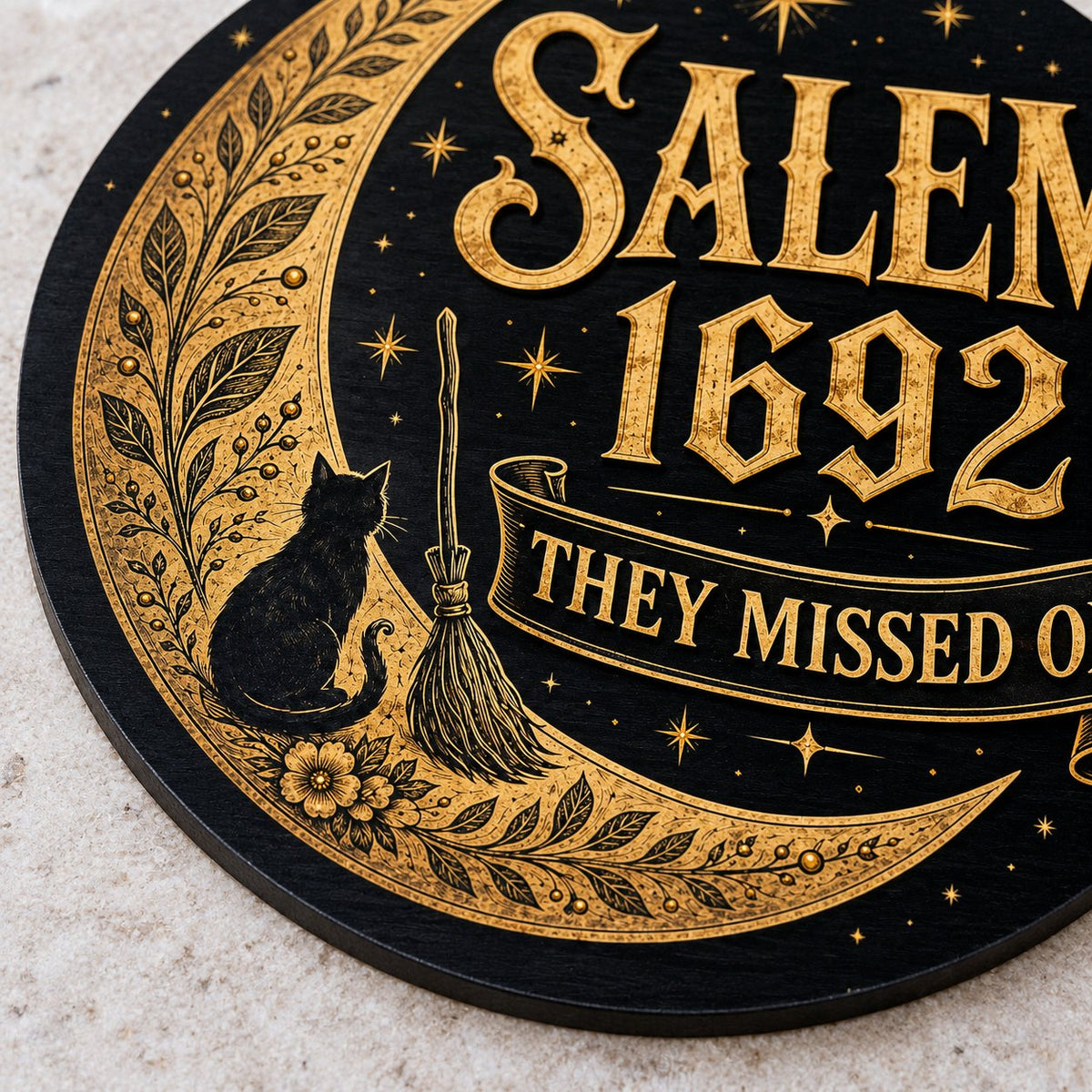 Round wooden sign with gold crescent moon, black cat, broomstick, stars and 'SALEM 1692 THEY MISSED ONE' on black.