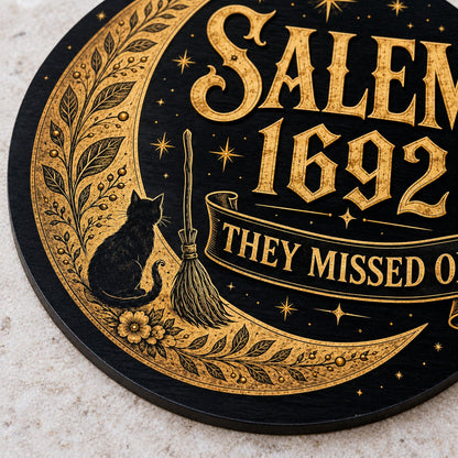 Round wooden sign with gold crescent moon, black cat, broomstick, stars and 'SALEM 1692 THEY MISSED ONE' on black.