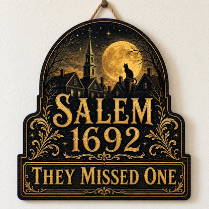 Custom-cut wooden sign reading “SALEM 1692” and “THEY MISSED ONE” with a black cat on a moonlit village and gold filigre