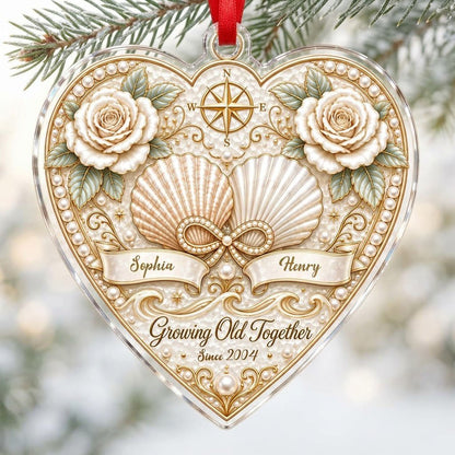 Heart-shaped clear acrylic ornament with seashells, pearl infinity symbol, compass star, and two name ribbons.
