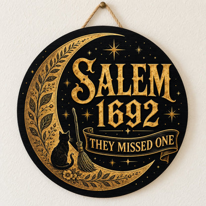 Round wooden sign with gold crescent moon, black cat, broomstick, stars and 'SALEM 1692 THEY MISSED ONE' on black.