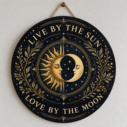 Round wooden sign with gold sun and crescent moon faces and the words “Live By The Sun, Love By The Moon”.