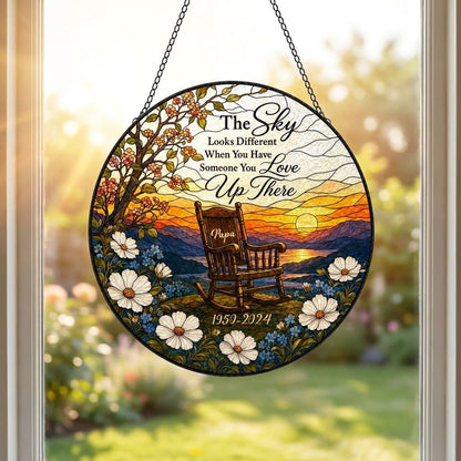 Round stained glass–style suncatcher with an empty rocking chair on a hill at sunset with flowers and a lake.