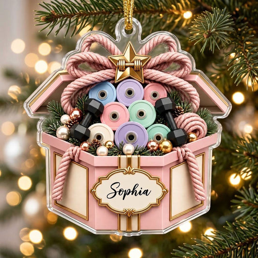 Pink gift box acrylic ornament with dumbbells, pastel weight plates, pine sprigs and a gold name label.