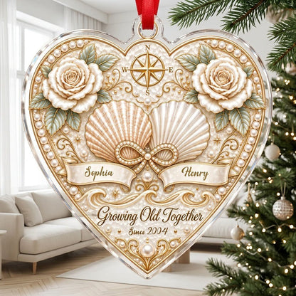 Heart-shaped clear acrylic ornament with seashells, pearl infinity symbol, compass star, and two name ribbons.