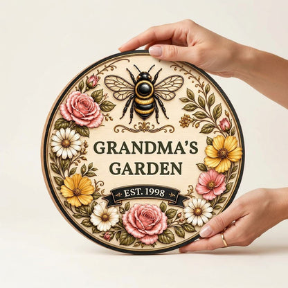 Round wooden sign with 'GRANDMA'S GARDEN', a gold-and-black bee, floral wreath, and EST. year on a light wood background