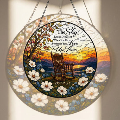 Round stained glass–style suncatcher with an empty rocking chair on a hill at sunset with flowers and a lake.