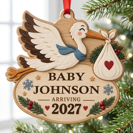 Custom-cut wood ornament of a flying stork with baby bundle above oval plaque reading BABY JOHNSON ARRIVING 2027.