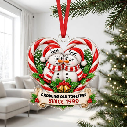 Two snowmen touching noses inside a candy-cane heart with holly, gold bells, and a scroll banner with names and year.