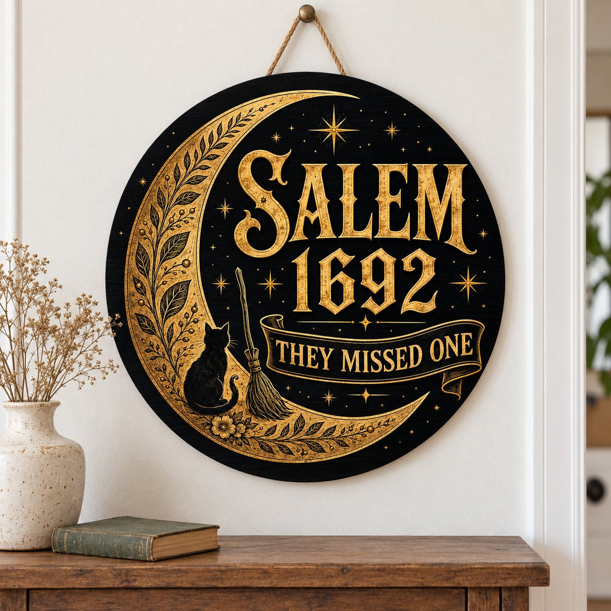 Round wooden sign with gold crescent moon, black cat, broomstick, stars and 'SALEM 1692 THEY MISSED ONE' on black.