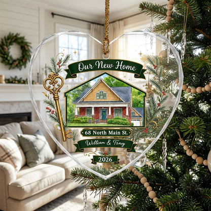 Heart-shaped glass ornament with watercolor house, gold key, evergreens, green banners, and 'Our New Home' text.