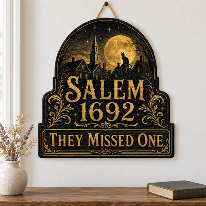 Custom-cut wooden sign reading “SALEM 1692” and “THEY MISSED ONE” with a black cat on a moonlit village and gold filigre