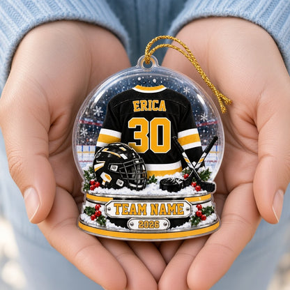 Snow globe acrylic ornament with black and gold hockey jersey, crossed sticks, puck, holly and a year plaque.