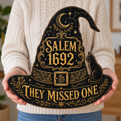 Witch hat-shaped wooden sign with 'SALEM 1692 THEY MISSED ONE', crescent moon, stars, filigree, and a black cat on the b