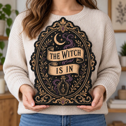 Gothic plaque sign reading "THE WITCH IS IN" with crescent moon, stars, purple smoke, potion bottle and ornate filigree 