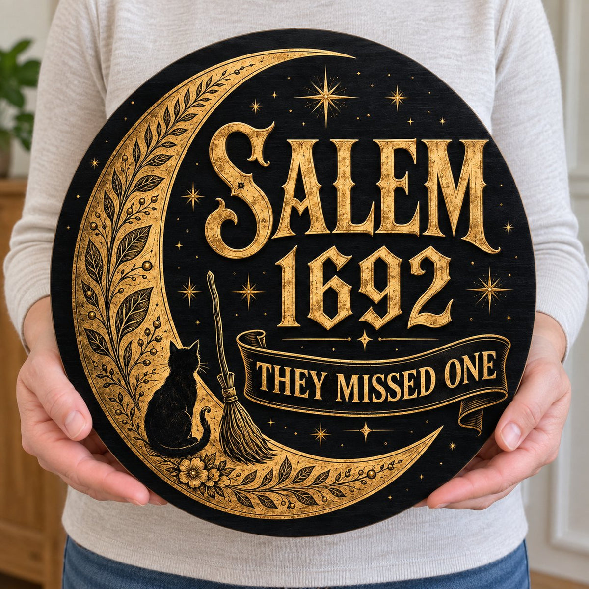 Round wooden sign with gold crescent moon, black cat, broomstick, stars and 'SALEM 1692 THEY MISSED ONE' on black.