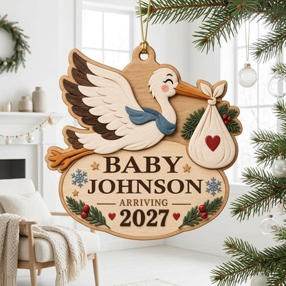Custom-cut wood ornament of a flying stork with baby bundle above oval plaque reading BABY JOHNSON ARRIVING 2027.