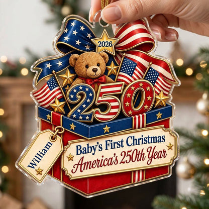 A teddy bear in a red gift box with blue trim, 250 and 2026 with American flags and a patriotic bow on an acrylic orname