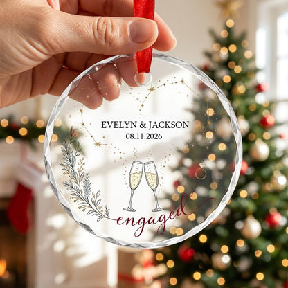 Round faceted glass ornament with gold constellation heart, ring, and clinking champagne flutes reading engaged.