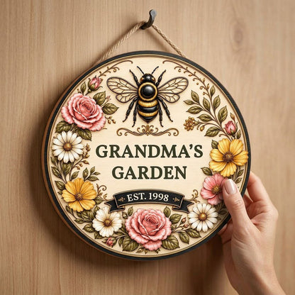 Round wooden sign with 'GRANDMA'S GARDEN', a gold-and-black bee, floral wreath, and EST. year on a light wood background
