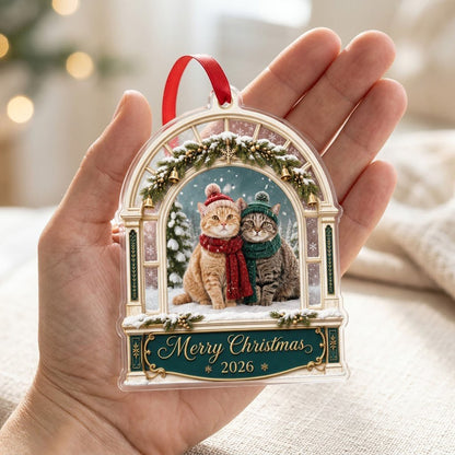 Arched window acrylic ornament with pine garland, gold bells, two cats in winter hats and scarves, and a green banner fo