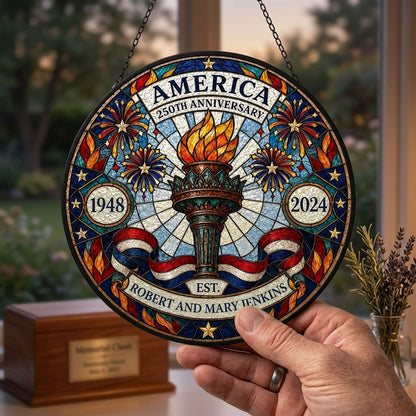 Round stained glass suncatcher with a central torch, fireworks, stars, and a red, white and blue ribbon.