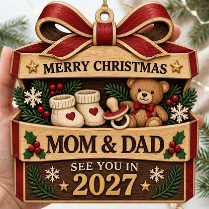 Gift box-shaped wood ornament with red bow, baby booties, teddy bear, and 'Mom & Dad See You In 2027' text.