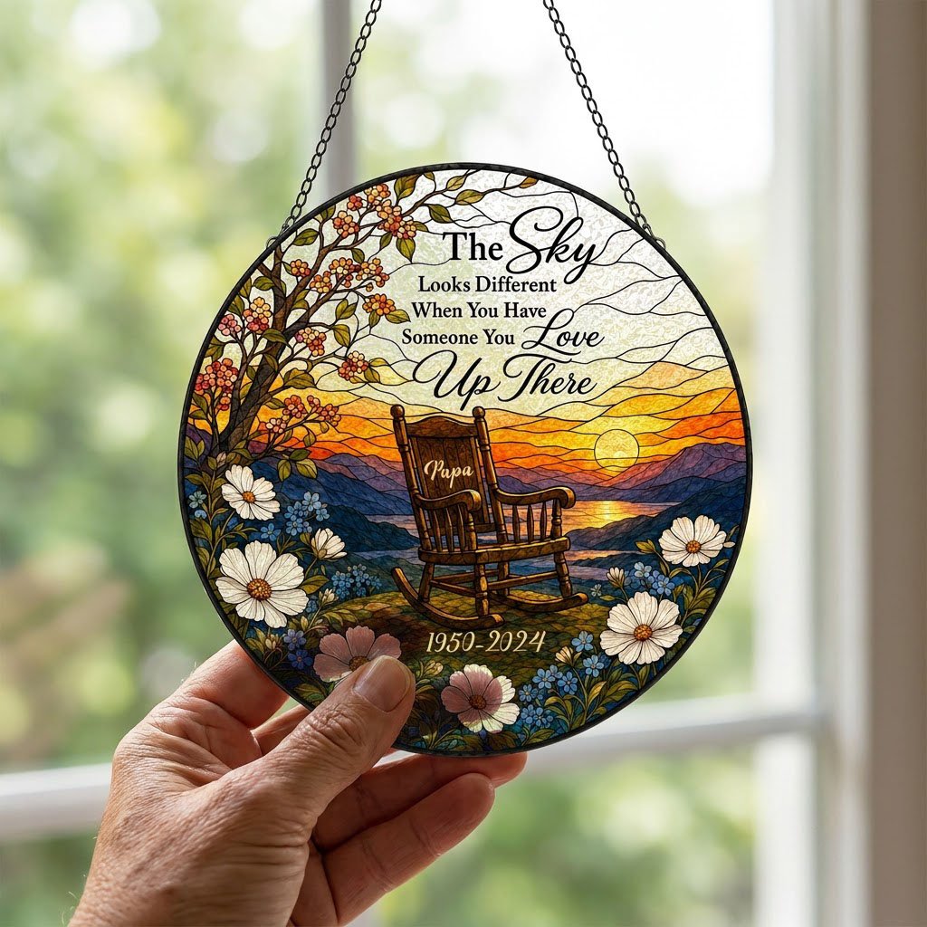 Round stained glass–style suncatcher with an empty rocking chair on a hill at sunset with flowers and a lake.