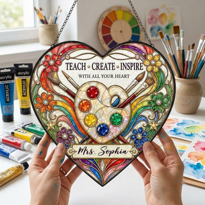 Heart-shaped stained glass suncatcher with palette, brushes, rainbow arcs, flowers and 'Teach • Create • Inspire'.